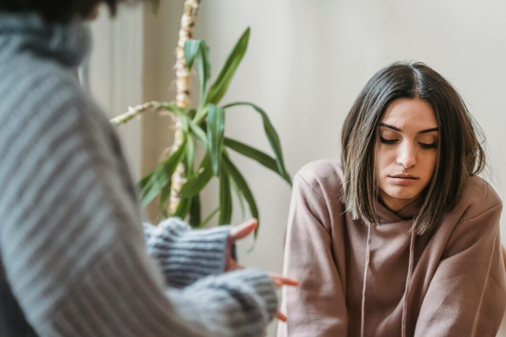 A woman with dark hair wearing a hoodie sits and looks downward during a conversation with another person.