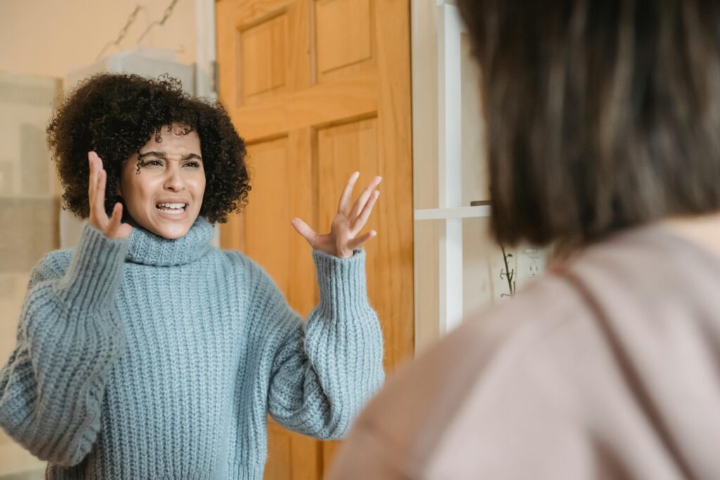 A woman with curly hair in a gray turtleneck sweater raises both hands while talking to another person standing at a doorway.