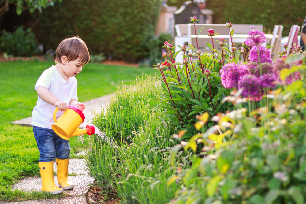 Cute little baby boy in yellow rainboots watering blooming flowers in garden from watering can. Summer gardening lifestyle. Little mommys helper.