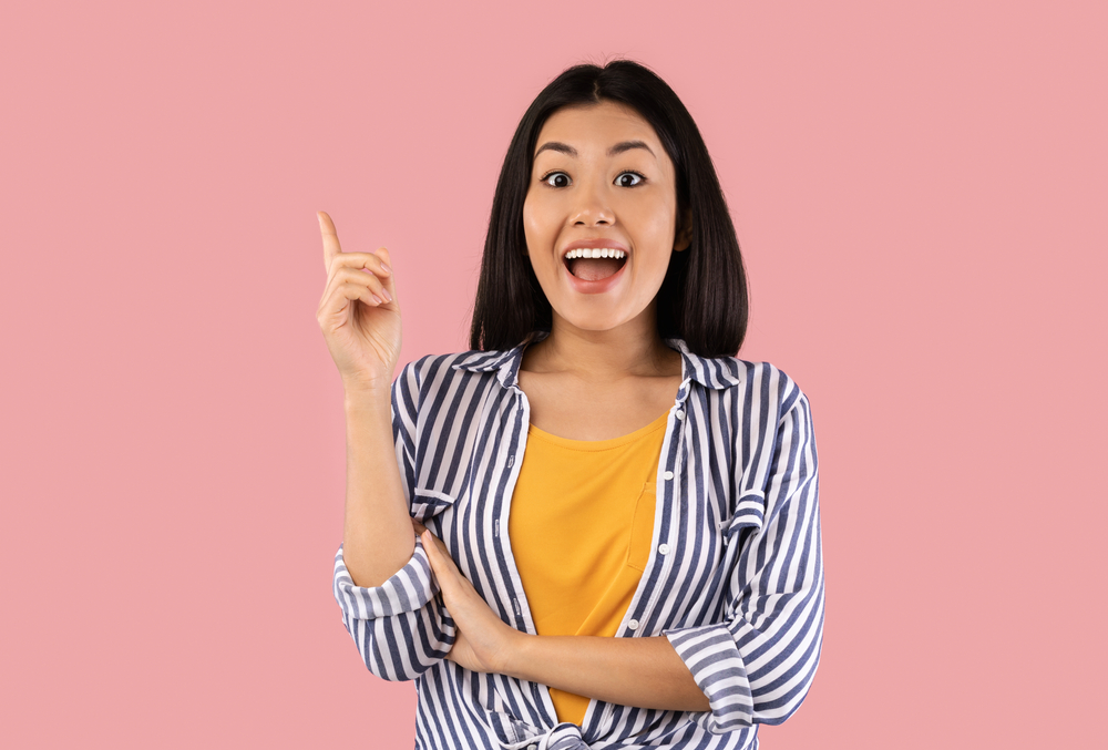 Aha Moment. Portrait of excited asian woman having great idea, finding inspiration or solution to problem. Cheerful young lady pointing finger up isolated over pastel pink studio background.