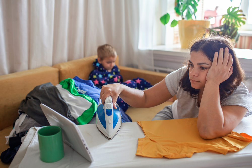 Tired middle aged mom watches video on tablet and ironing things at the same time, next to her son with smartphone too, social media and gadget addiction, housewife and mom burnout concept