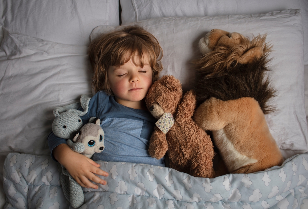 Healthy child, sweetest blonde toddler boy sleeping in a bed with a teddy bear and another stuffed animals. Beautiful baby boy have a healthy sleep in the bed. Sleeping baby.