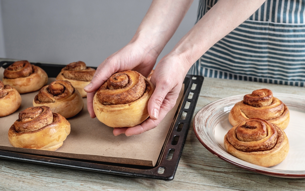 Woman in a blue apron is taking out fresh warm cinnamon rolls buns from a baking tray and putting them on a plate. Concept of delicious homemade pastries and cozy atmosphere.