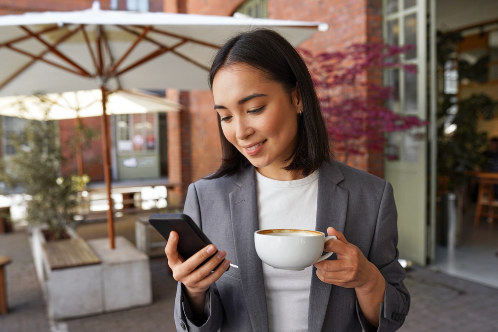 Young elegant Asian professional business woman, lady executive manager wearing suit holding coffee cup standing outdoor holding smartphone using mobile apps on cell phone during work break.