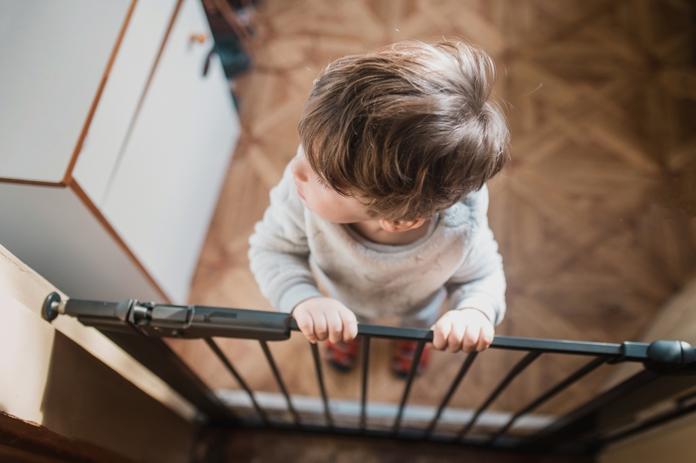 little boy standing on the other side of the child safety gate and holding on to the top of the security gate. Alone at home without parental advisory Selected focus