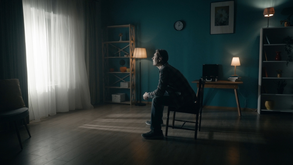 A man sits on a chair in the middle of the room and looks at the light in the window. Side view of a lonely man sitting in a dark room. Hope concept, mental health.