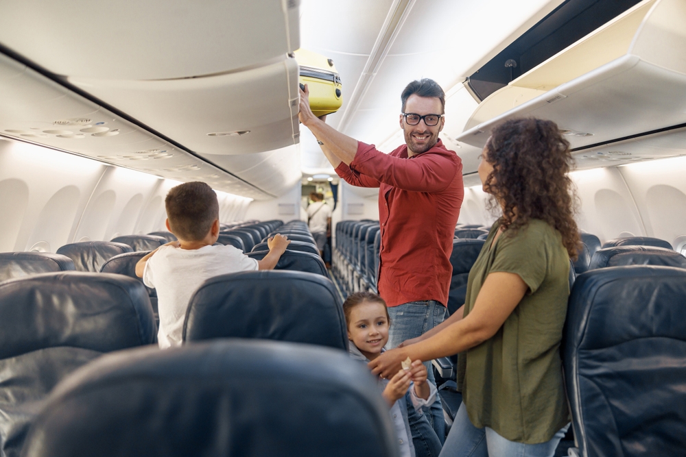 Cheerful man looking at his wife with a smile and putting carry on luggage in compartment while traveling together with his family by plane. Family vacation, transportation concept