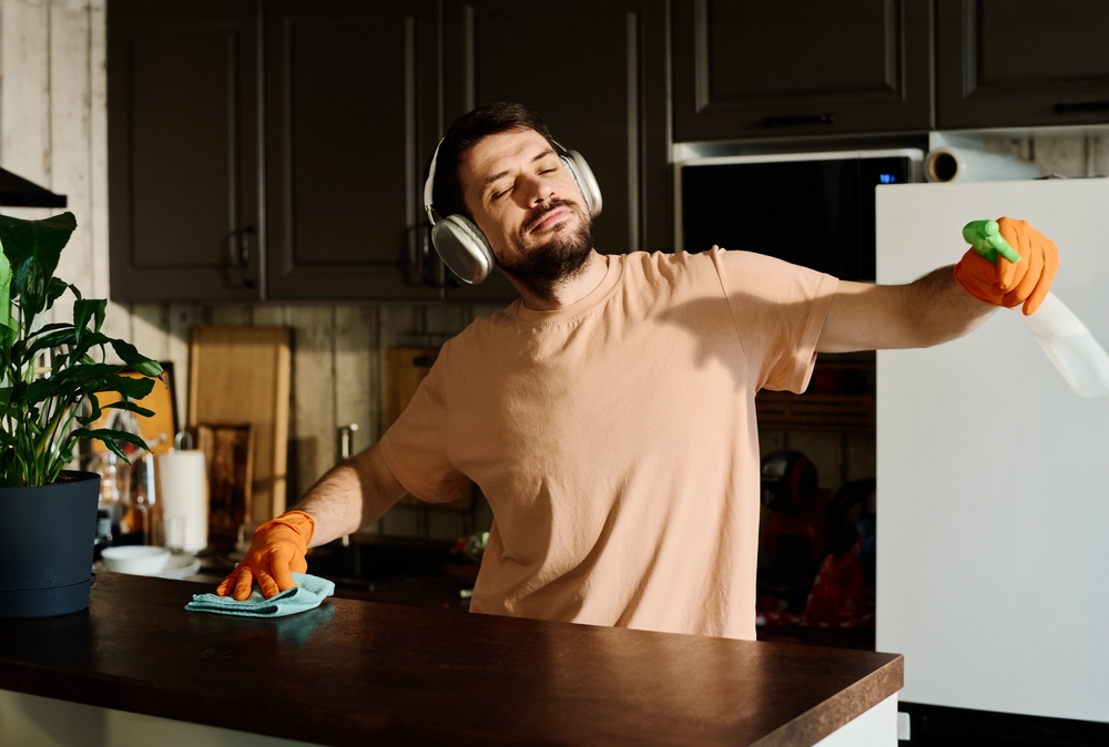 Young man in headphones enjoying sunny day and his favorite music while standing by kitchen counter and carrying out domestic chores