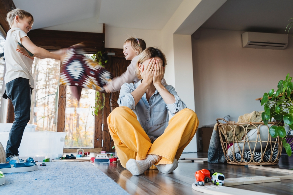 Stressed out mother sitting on floor in middle of toys while children naughty running around her at room. Frustrating mom stressful feels helpless struggling with problems while kids playing around