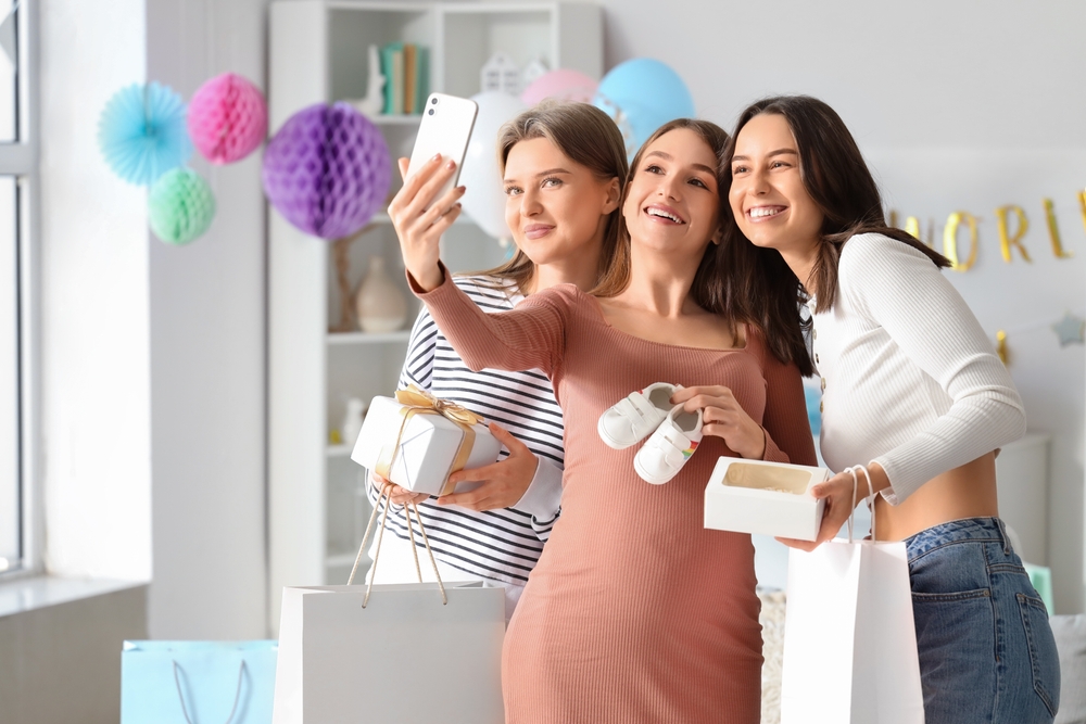Young pregnant woman with her friends and gifts taking selfie at baby shower party