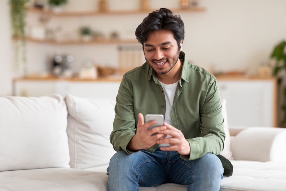 Arab man is seated comfortably on a couch, engrossed in his phone screen. The room is dimly lit, emphasizing his focused expression as he scrolls through content on his device.