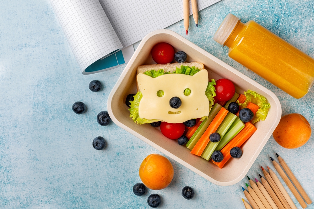 Back to school background. Kids school lunch box with funny fox sandwich with cheese, and fresh vegetables, fruits, orange juice. Colorful pencils, copybook. Blue background, copy space.