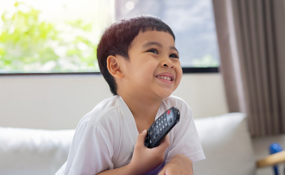 Relaxed cute Asian little cute boy sitting on couch holding remote controller, watching TV at home, enjoying his free time at weekend, copy space
