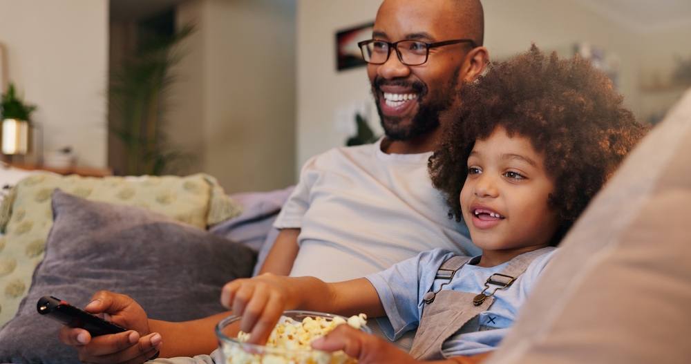 Father, boy and happy with television or popcorn on sofa in living room for movies, cartoon and entertainment. People, parent and smile with kid at home for streaming platform with child development