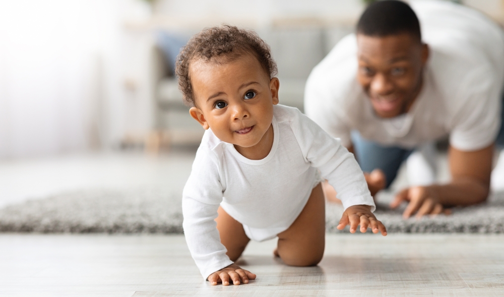 Cute Little Black Infant Baby Crawling On Floor At Home, Proud Young Father Looking At Him And Smiling, Dad And Toddler Child Enjoying Spending Time Together, Selective Focus With Copy Space