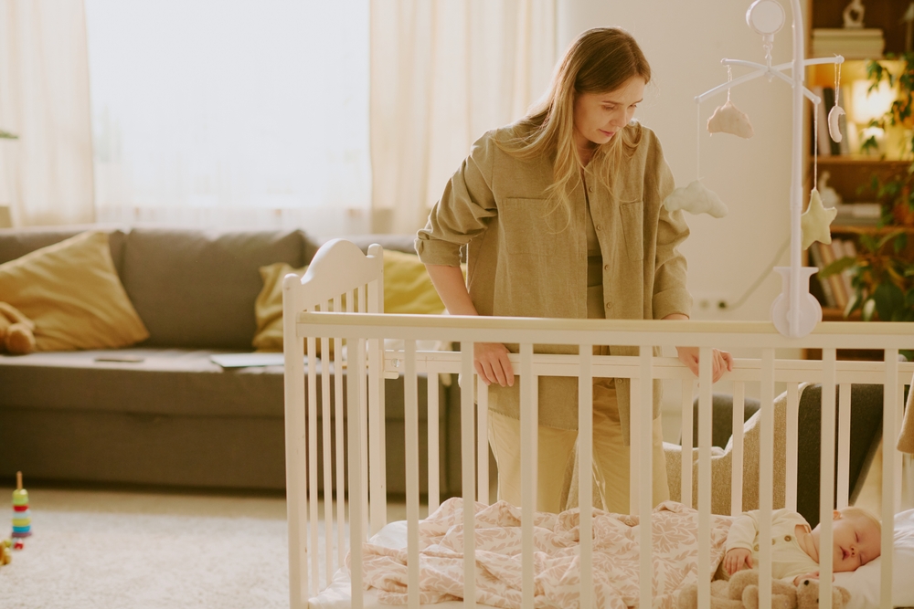 Mother standing by crib watching her sleeping baby. Soft beige decor and sunlight from large windows create a warm and serene ambiance in living room