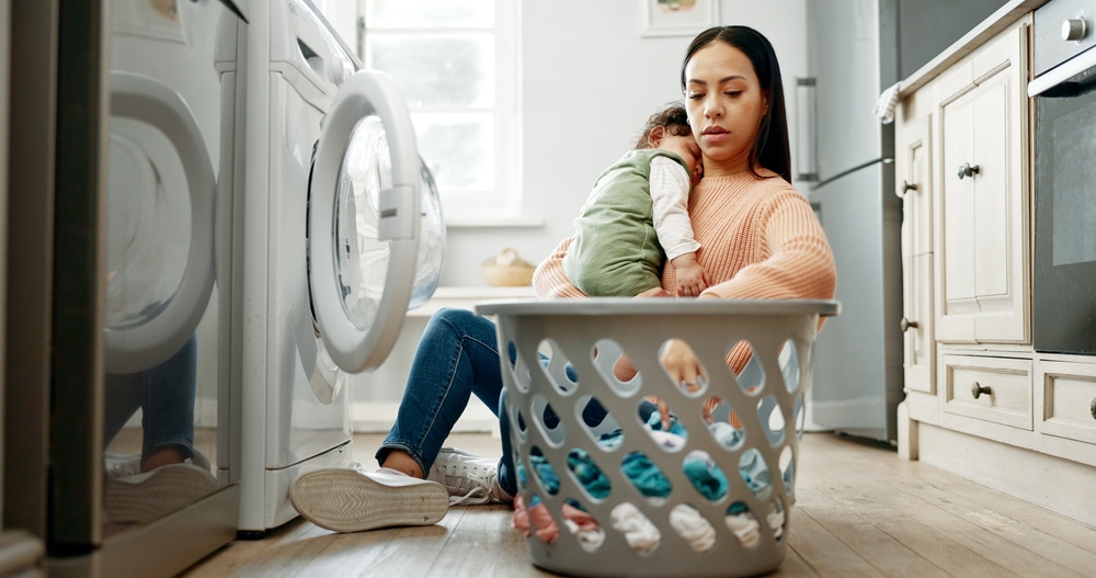 Mother, baby and basket in kitchen for laundry, housekeeping and motherhood in house with sleeping child. Woman, newborn and tired with washing machine for cleaning clothes, parenthood and burnout