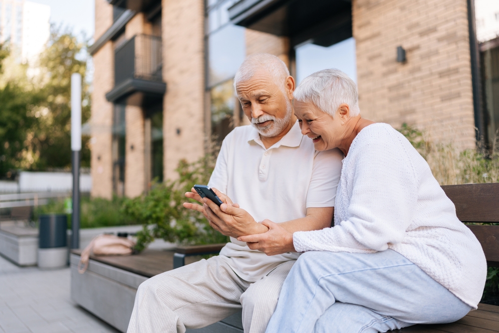 Positive senior couple enjoying time sitting hugging in city bench, happily using smartphone, browsing internet together. Happy gray-haired male and female looking at something funny on smartphone.