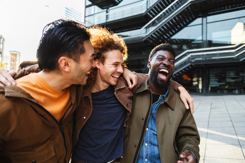 Three young male friends are walking together outdoors in an urban setting, arms around each other, laughing and enjoying each other's company