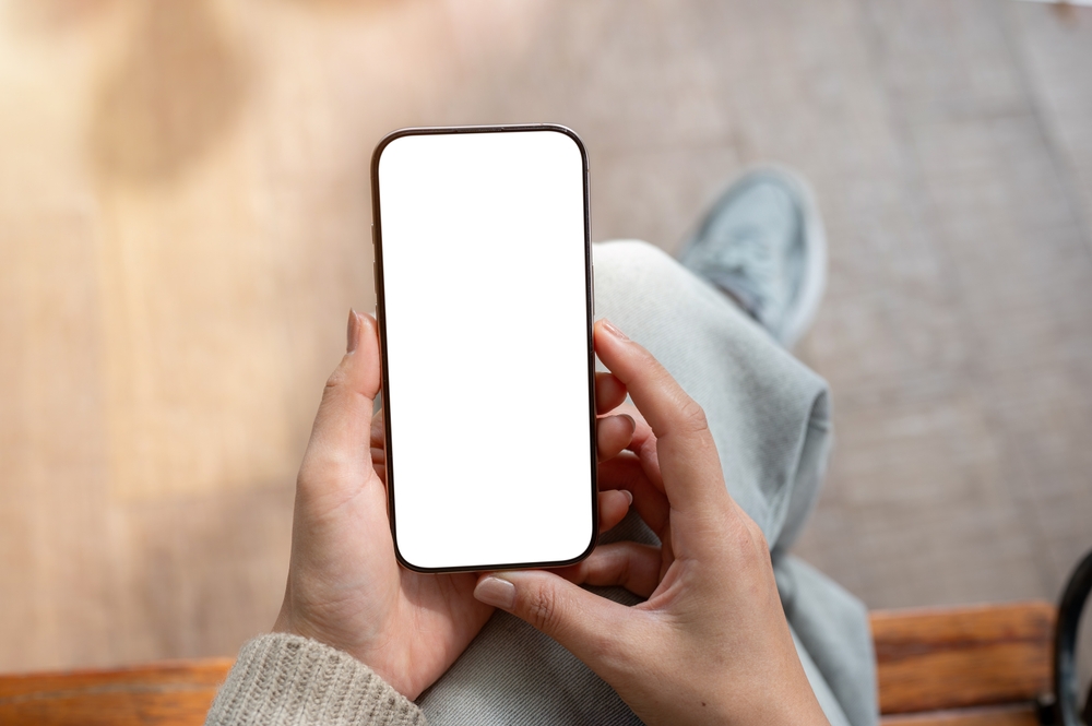A top view of a woman sits indoors and holds a smartphone with a white screen mockup, with blurred background. people and wireless technology concepts