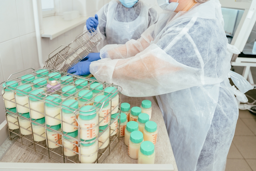 Two lab workers in protective wear handling donor breast milk containers and organizing them into metal baskets for further processing in milk bank environment.