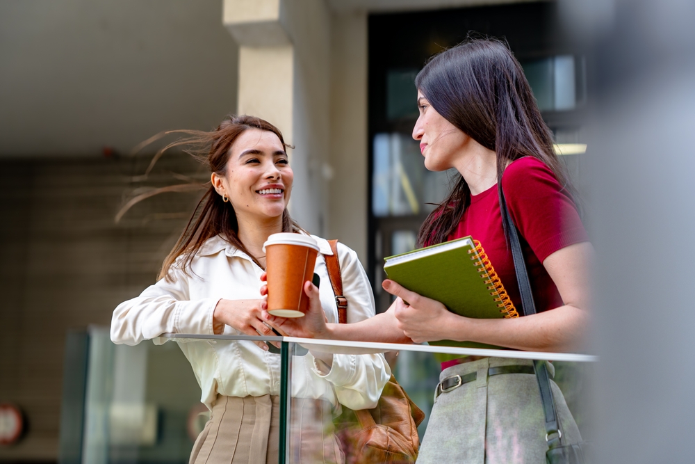 Two female colleagues enjoying a coffee break, talking and smiling outside the office building