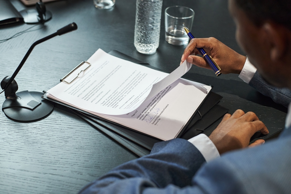 Middle aged Black man reviewing official documents at desk, holding pen and turning page, sitting beside microphone and water bottle, participating in political meeting