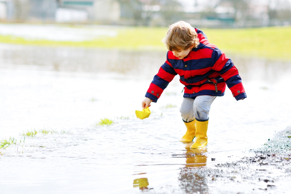 Happy little kid boy in yellow rain boots playing with paper ship boat by huge puddle on spring or autumn day