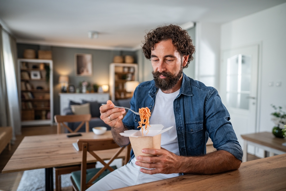 Bearded man in denim shirt eating takeout noodles from a carton at a wooden table in his modern apartment, relaxed and enjoying a quick lunch or dinner at home