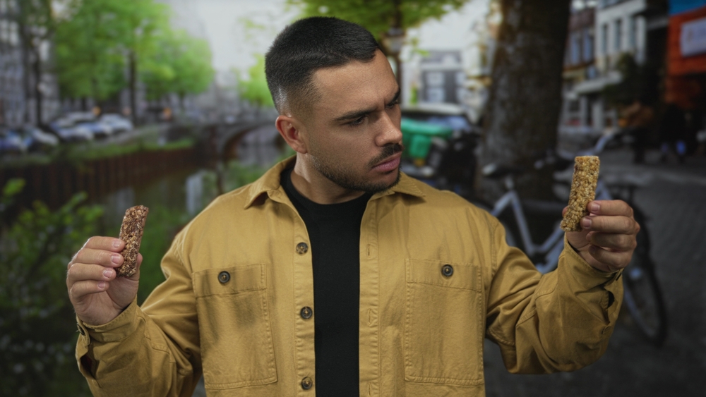 Man holding two crunchy granola bars grimaces by canal street in the city as he inspects texture and aroma; disgust.