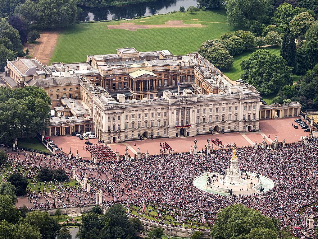 Aerial view of Buckingham Palace with large crowds gathered on the grounds and surrounding streets.