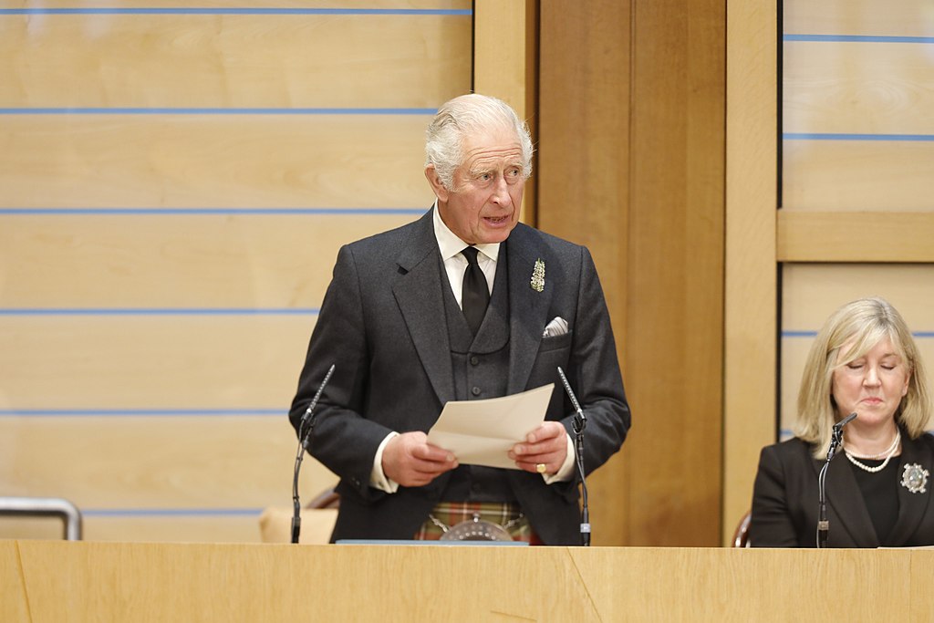 Charles in a dark suit and tartan kilt speaking at a podium in the Scottish Parliament chamber.