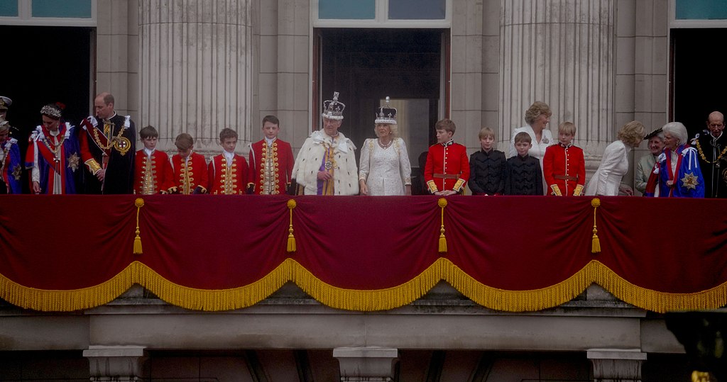 Members of the royal family standing on the Buckingham Palace balcony draped in red velvet, with several wearing coronation robes and page uniforms.
