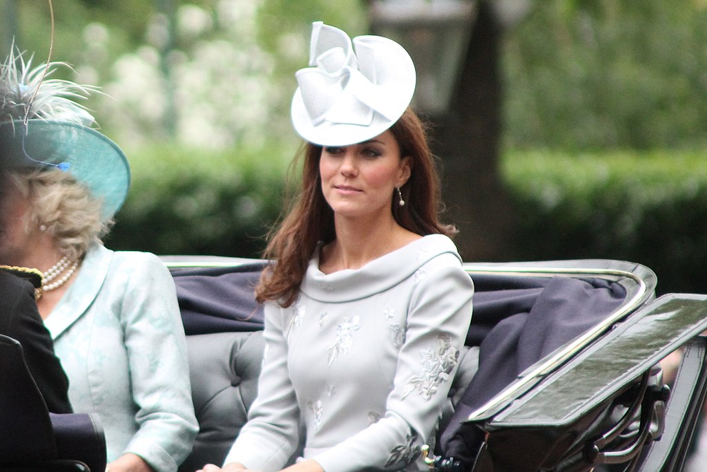 Kate Middleton in a pale grey coat and white decorative hat riding in an open carriage.
