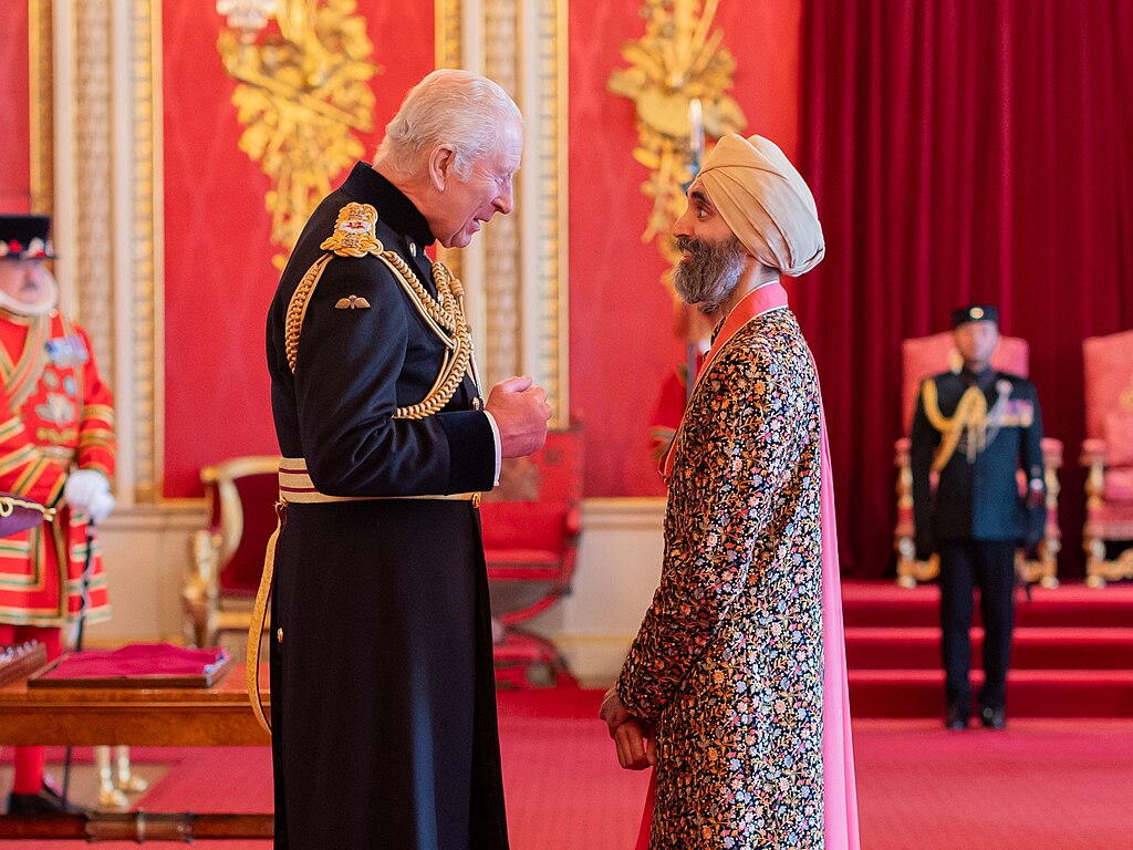 King Charles III in dark military uniform and Jasvir Singh CBE wearing a turban and embroidered coat in conversation at a Buckingham Palace ceremony.