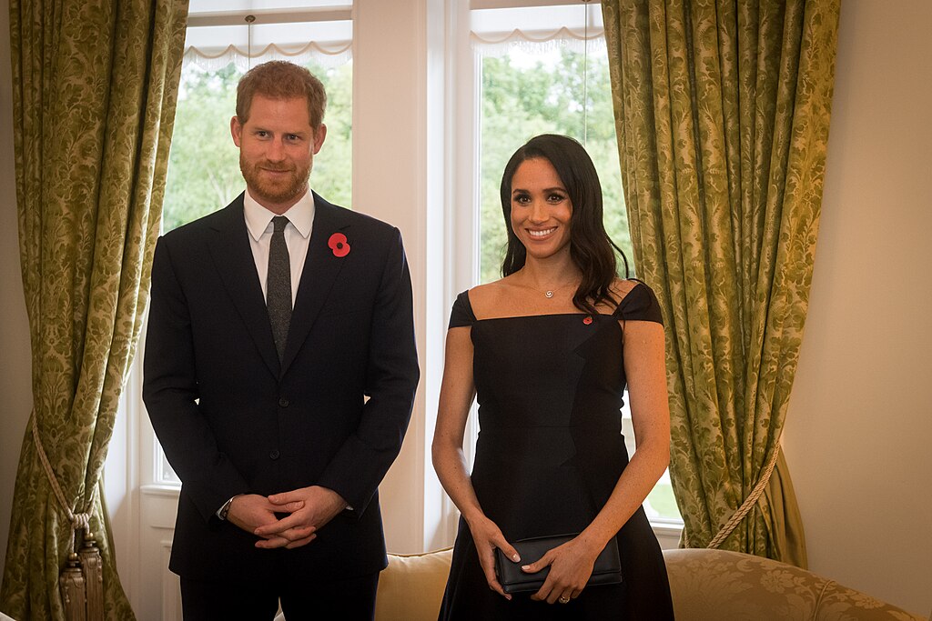 Prince Harry in a dark suit with a poppy pin and Meghan Markle in a black off-shoulder dress standing in a formal room.