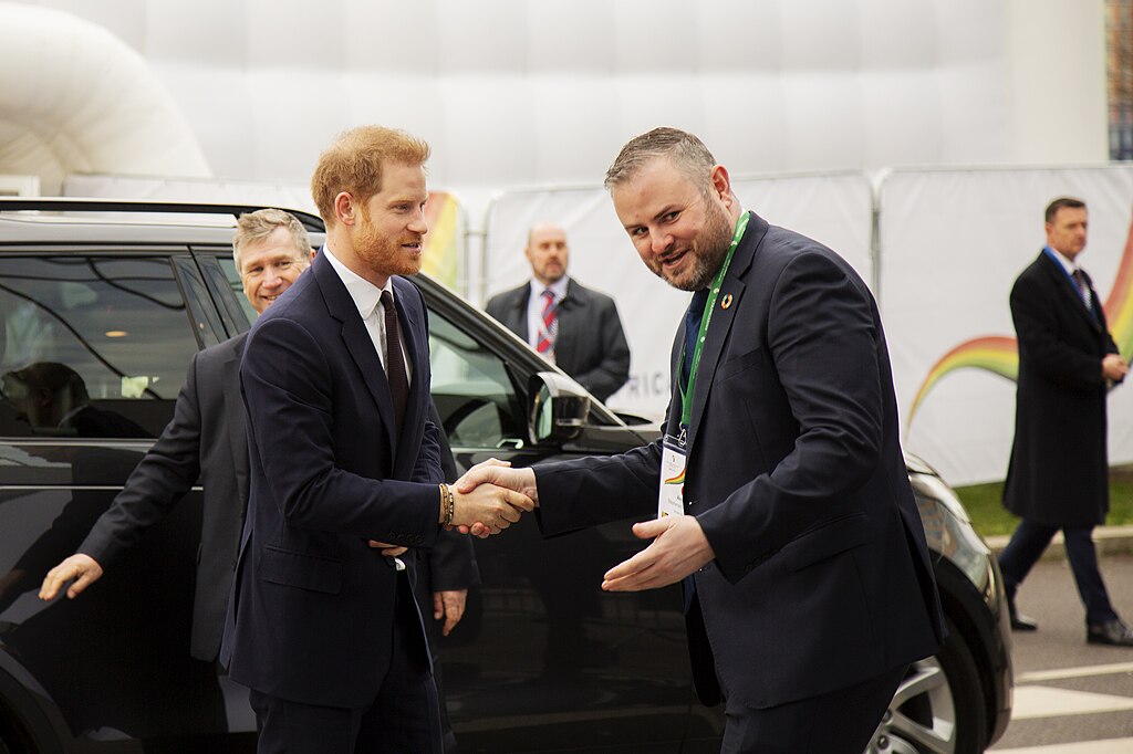 Prince Harry in a dark suit shaking hands with a man outside an event venue.