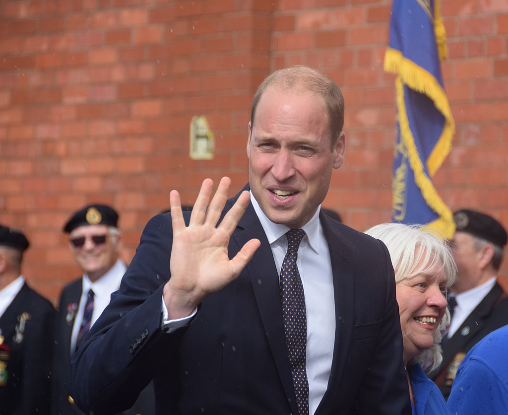 Prince William in a navy suit waving to a crowd, with veterans visible in the background.