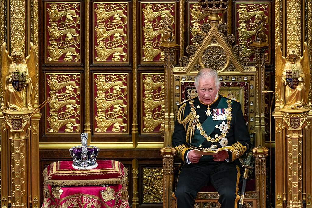 King Charles III in green military dress uniform reading from papers while seated on a gilded throne, with the Imperial State Crown displayed on a table beside him.