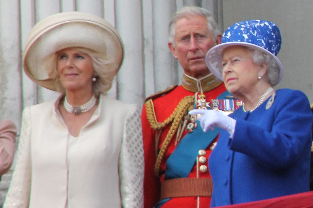 Camilla in a cream outfit and hat, Charles in red military uniform, and Queen Elizabeth II in a royal blue coat and hat standing on a balcony.