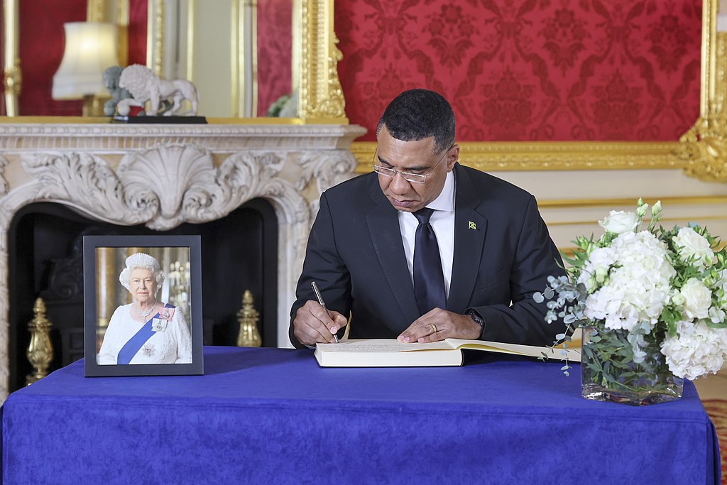 Jamaican Prime Minister Andrew Holness in a dark suit signing a condolence book at a table draped in blue cloth, with a framed portrait of Queen Elizabeth II and white flowers nearby.