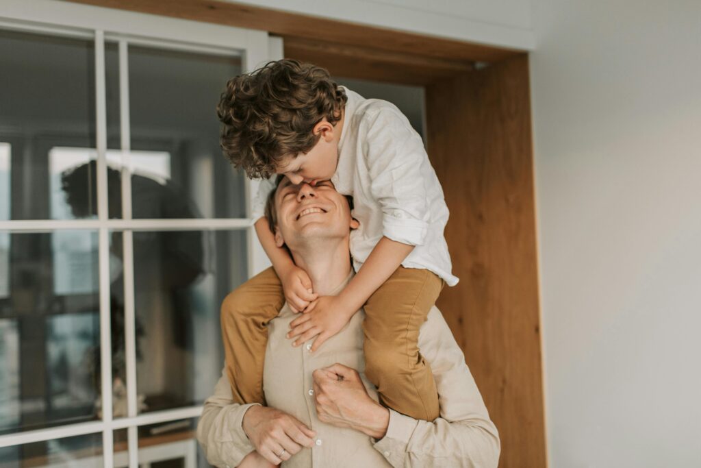 A young boy with curly hair sits on his father's shoulders, leaning down to kiss his forehead. The father smiles broadly, eyes closed, arms wrapped around the child's legs.