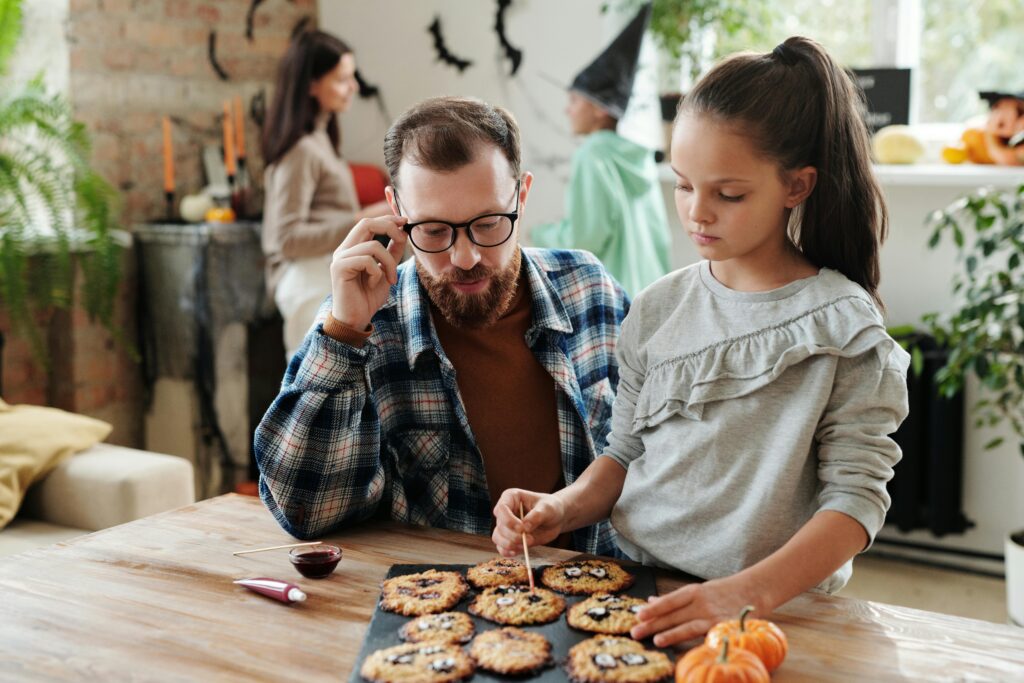 A bearded father in glasses and a plaid shirt decorates Halloween cookies with his young daughter while talking on the phone. The mother stands blurred in the background amid Halloween decorations including paper bats.