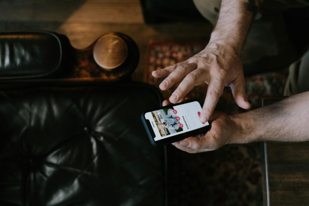 Overhead view of a man's hands scrolling through a dating app on his phone while seated on a dark leather couch.
