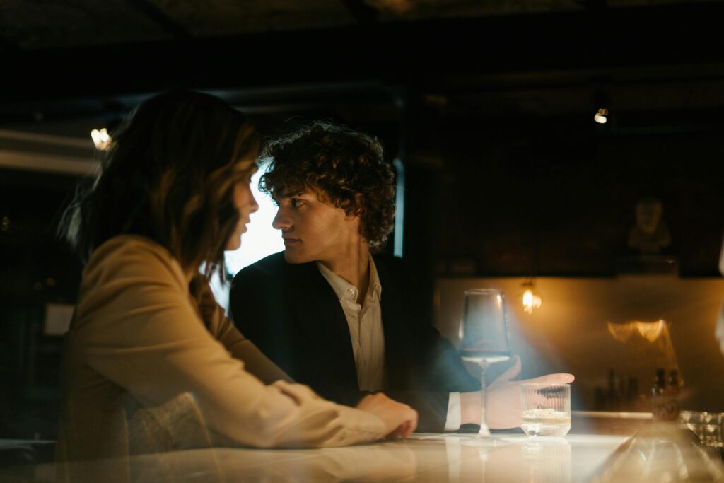 A couple sits close together at a dimly lit bar, but the man with curly hair glances suspiciously around the room rather than focusing on his companion. Glasses sit on the counter before them.