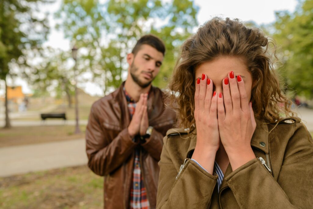 Outdoors, a woman with curly hair and red nail polish covers her face with both hands while a man in a brown leather jacket stands behind her with hands pressed together pleadingly.