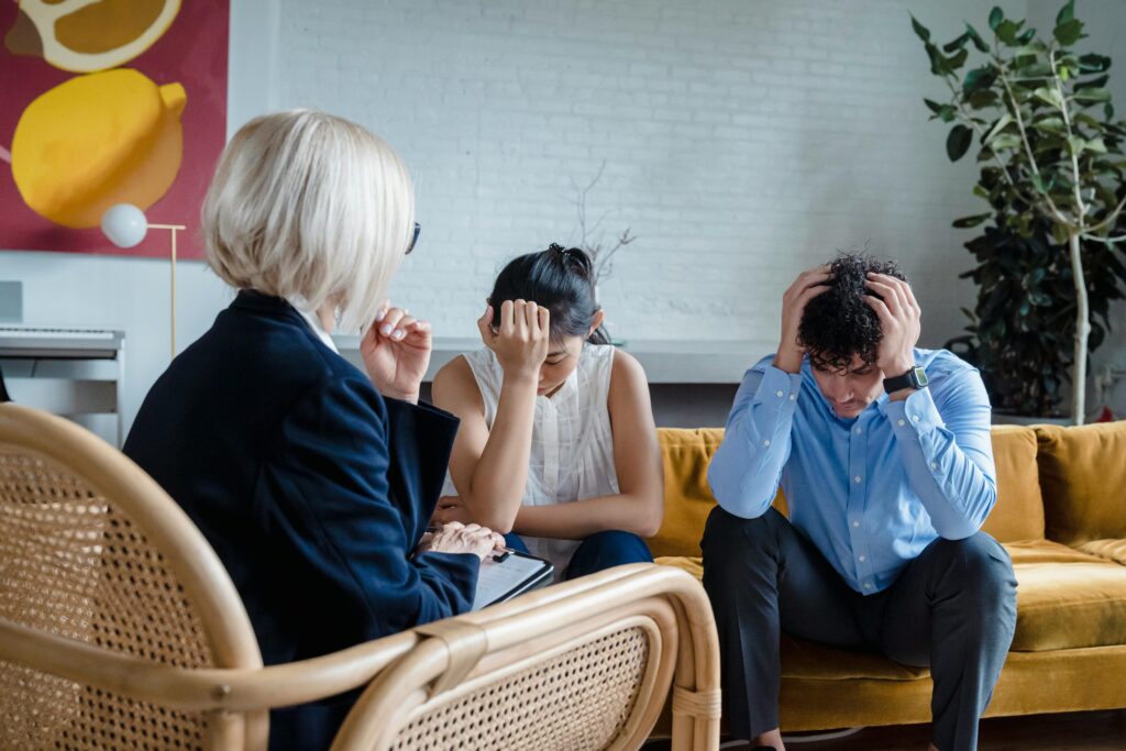 A couple sits on a mustard-yellow couch during therapy, both with heads in their hands in distress, while a therapist with short white hair in a navy blazer sits across from them taking notes.