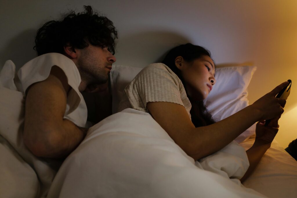 A couple lies in bed with warm amber lighting. The man with dark curly hair looks toward the woman, who faces away from him while scrolling on her phone.