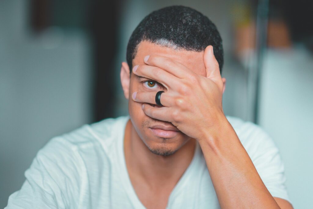A man peers through his fingers while covering his face with one hand, wearing a white v-neck shirt and dark ring.