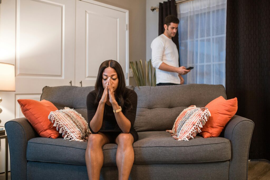A woman with long dark hair sits on a gray couch with coral pillows, hands pressed together in front of her face in distress. Behind her, a man in a white shirt stands looking at his phone, emotionally disconnected.